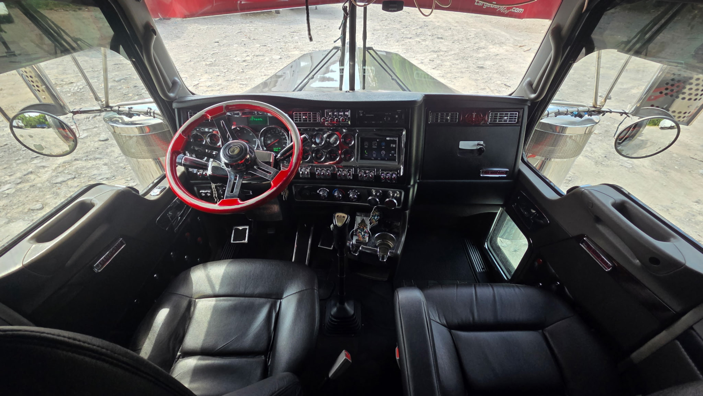 Kenworth W900L cab interior showing red steering wheel, dashboard gauges, and black leather seats viewed from passenger side with large windshield and mirrors