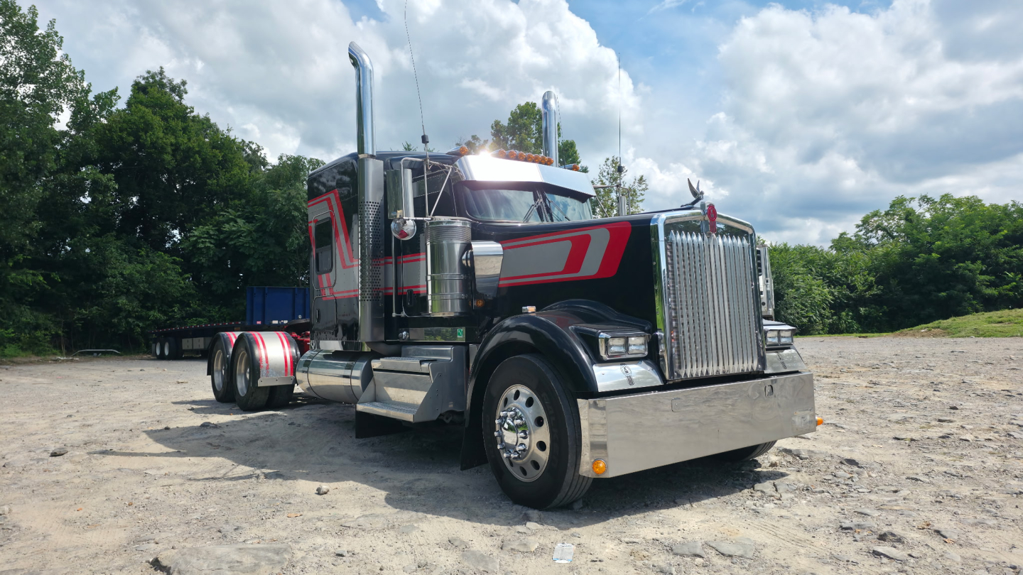 Kenworth W900L truck in a parking lot, front three-quarter view, chrome grille, large exhaust stacks, black and red livery, aluminum fuel tanks, dual rear axles.