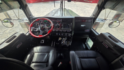 Kenworth W900L cab interior showing red steering wheel, dashboard gauges, and black leather seats viewed from passenger side with large windshield and mirrors