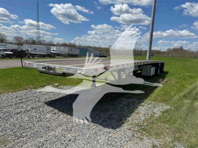 Flatbed trailer showing a long aluminum deck with low profile, side rails, and front gooseneck area. Clear sky, grassy lot, other trailers in background.
