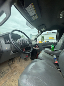 Interior of Ford F-750 dump truck cab showing steering wheel, dashboard, sun visor, and two seats with beverage bottles; dirt on floor and gear shifter visible.