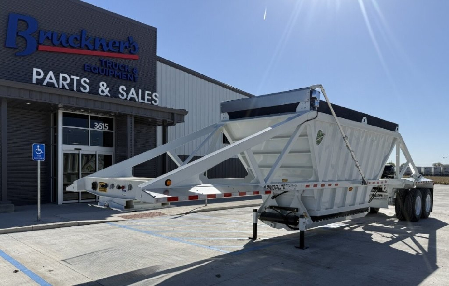 Trailer, Armor Lite SBD40 skeleton dump trailer in front of parts & sales building showing gooseneck hitch and dual-axle rear with white frame.