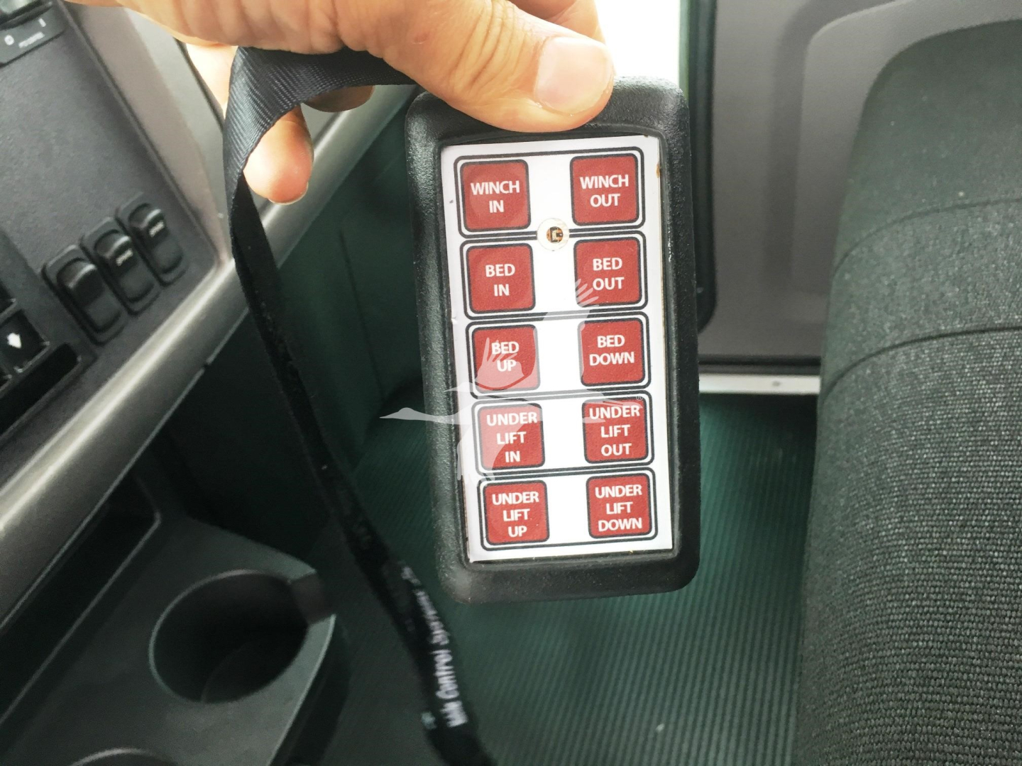 Trucker's handheld control box with red labeled buttons near seat area. Buttons include WINCH IN/OUT, BED IN/OUT, BED DOWN/UP, UNDER LIFT IN/OUT, UNDER LIFT UP, etc.