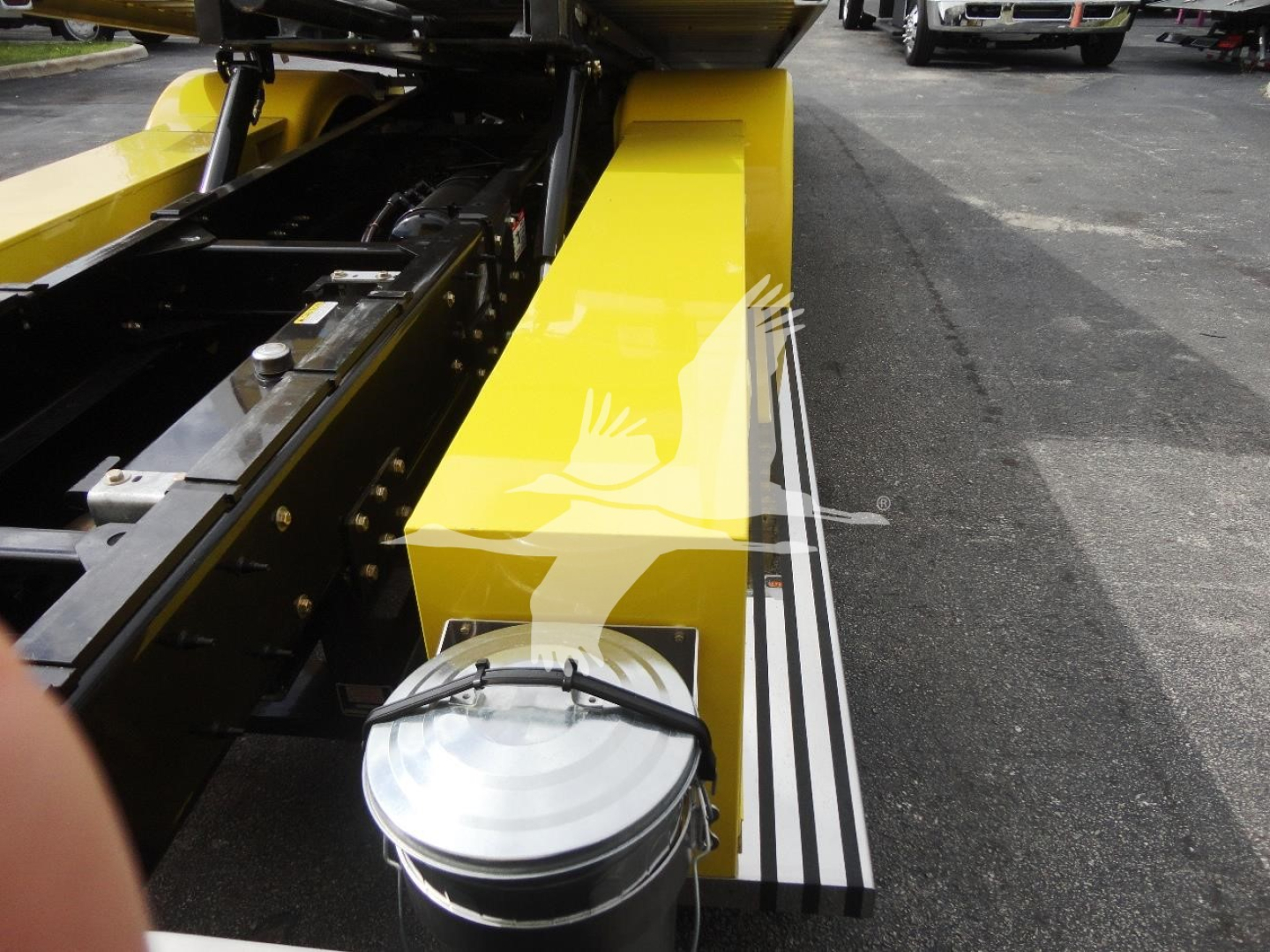 Truck component close-up showing a bright yellow side panel and mechanical linkage of a rollback tow truck, with a white circular cap on a metal tank and black rails.