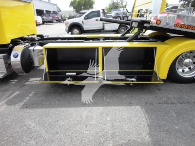 Truck rollback bed mounted on a 2022 Peterbilt 337 with bright yellow paint and hydraulic wings visible, adjacent equipment and other trucks in the lot.