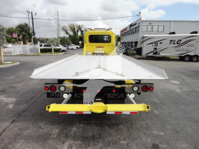 Rear view of a yellow Peterbilt 337 rollback tow truck with a large aluminum flatbed and rear lights, in a dealership lot.