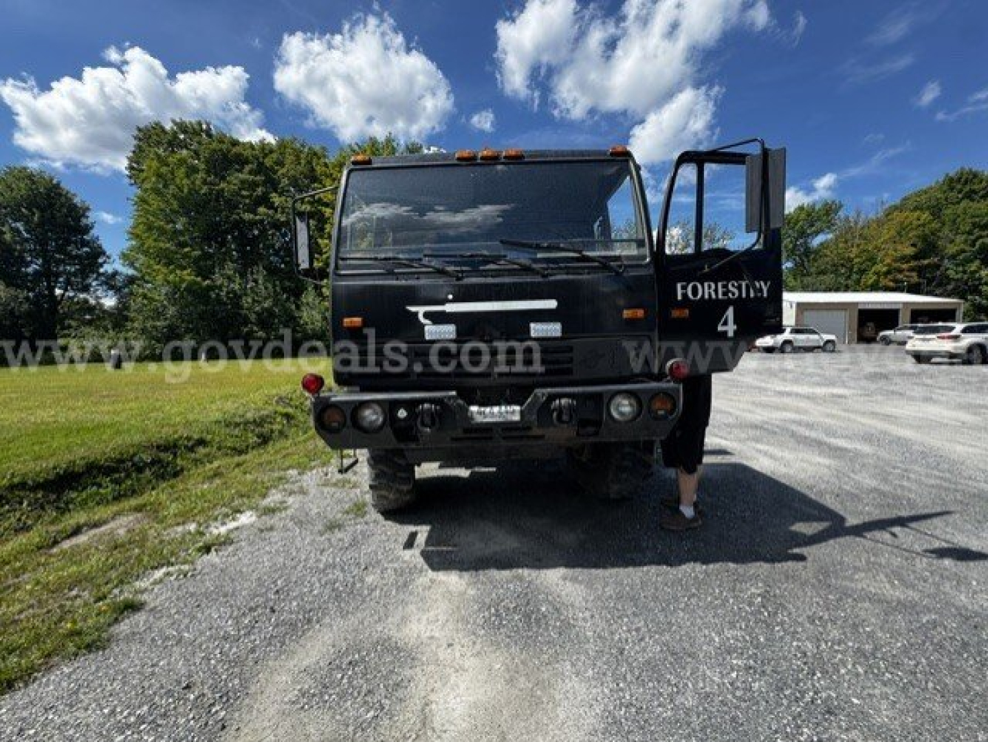 Truck front view with open passenger door showing 'FORESTY' text on the side, headlights, bumper, and grille; Muddy gravel lot background with trees.