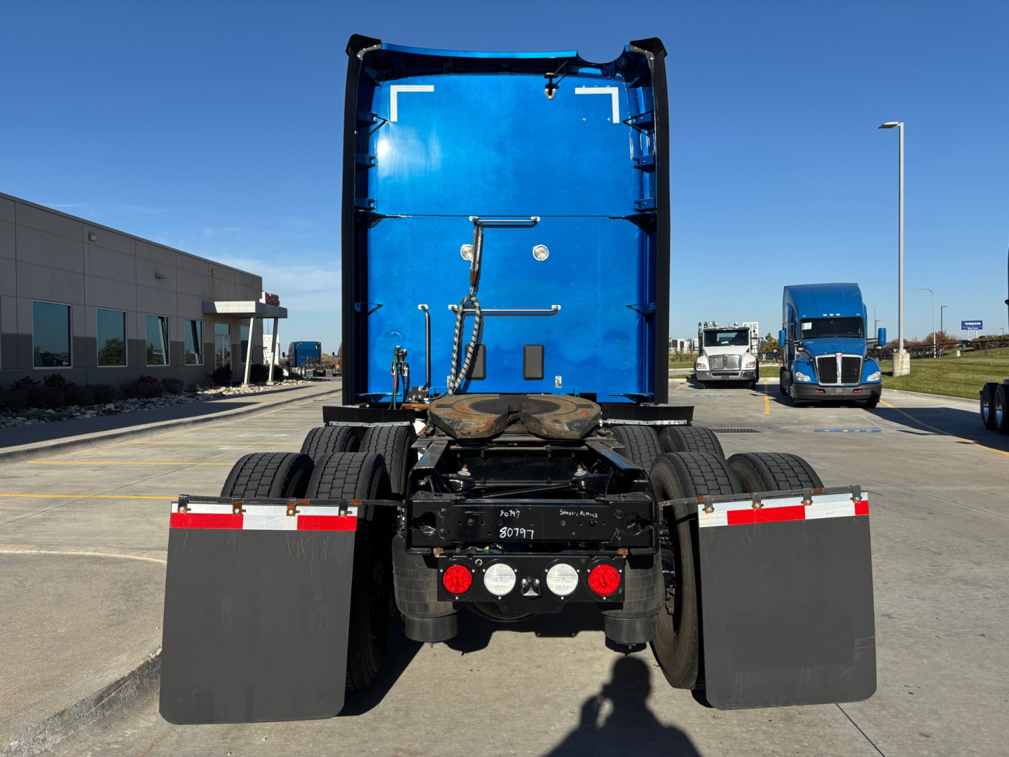Kenworth T680 rear view showing chassis, fuel/air lines, rear lights, and mud flaps with blue sleeper cab in background.