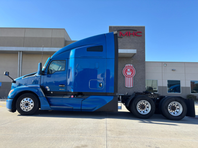 Kenworth T680 conventional sleeper tractor in blue, side view, showing cab, exhaust, and dual rear axles in front of a dealership building.