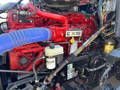 Kenworth T680 engine bay showing red  X15 branding on valve cover, blue intake hose, white Accar brake fluid reservoir, wiring, and various hoses.