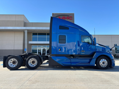 Blue 2023 Kenworth T680 tractor-trailer side view showing cab with windows and multiple wheels, parked in front of a building.