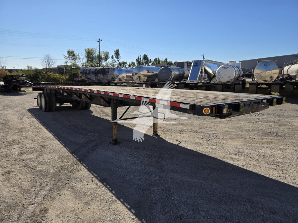Flatbed trailer in yard with multiple tanks in background; Fontaine brand flatbed trailer with long deck and supporting legs visible.