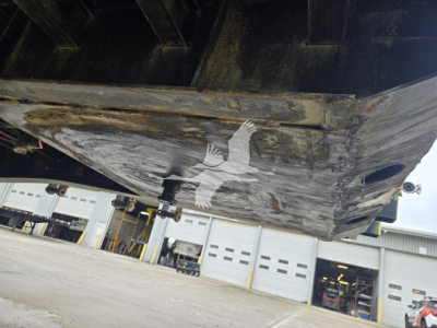 Flatbed trailer underside with visible metal frame and lower edge; a watermark of a bird overlay near center. No legible serials or labels visible on the trailer in this shot.