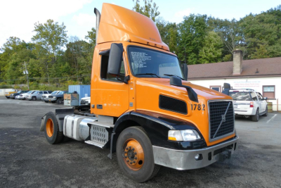 Truck cab in orange and black, Volvo VNM42T daycab, front three-quarter view, fuel tanks and steps visible, dealership lot in background.