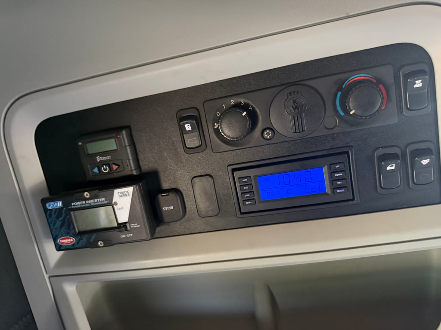 Kenworth T680 dash showing control panel with blue digital display, knobs, and switches. Seats or dashboard area visible.