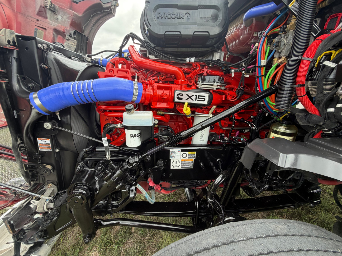 Kenworth T680 engine bay with red engine, blue intercooler hose, white reservoir, and exposed wiring harnesses.