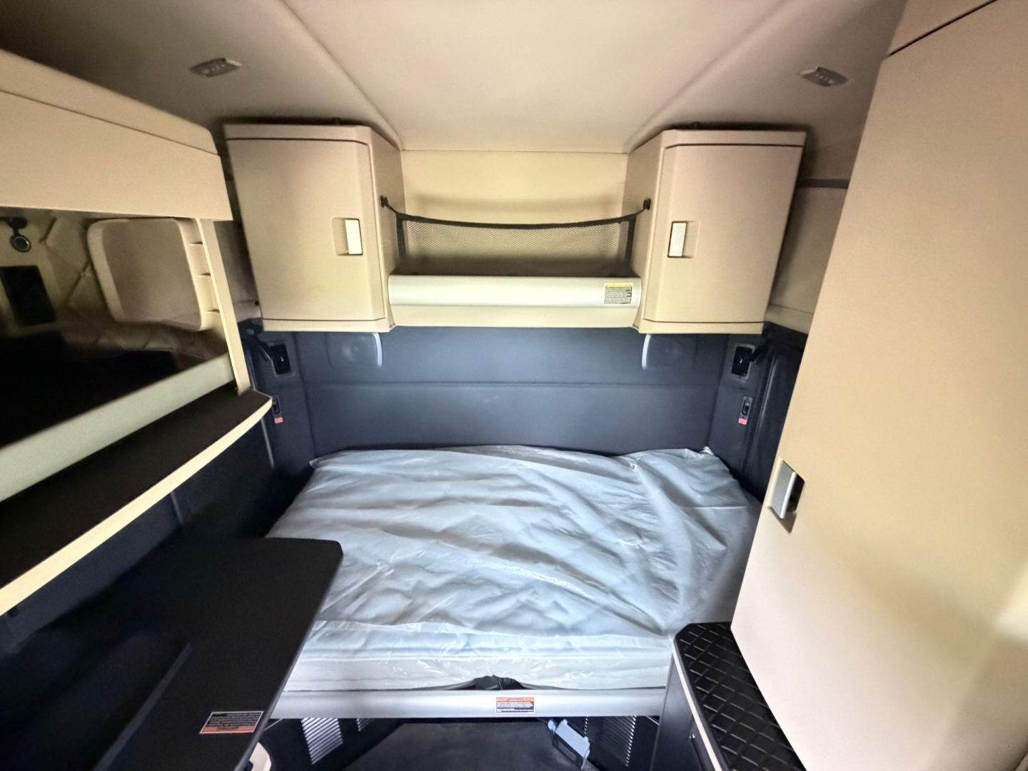 Cab interior of a Kenworth T680 sleeper showing a raised bunk bed with light gray bedding, overhead cabinets, and side shelves.