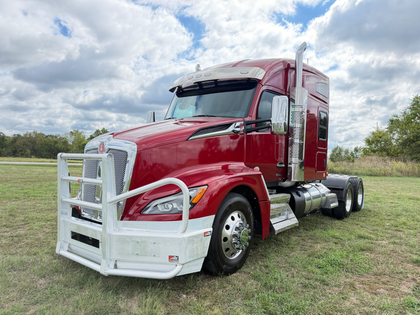 Kenworth T680 tractor unit in red with tall sleeper cab and chrome front grille guard, side exhaust, and dual rear axles on grass field.