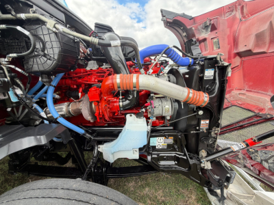 Kenworth T680 engine bay showing red engine block, blue hoses, black radiator support, and exposed components under the hood.
