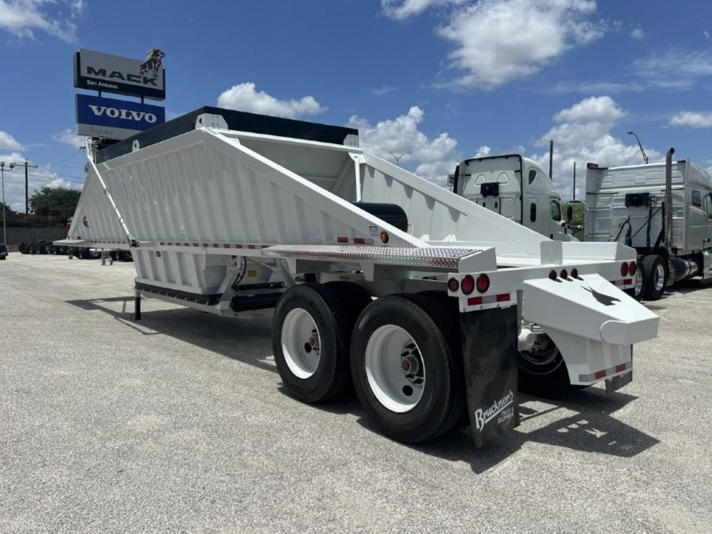 Dump trailer with white steel body and tandem axles, viewed from rear-left. Brand Brakes and mudflaps visible, dump bed raised slightly.