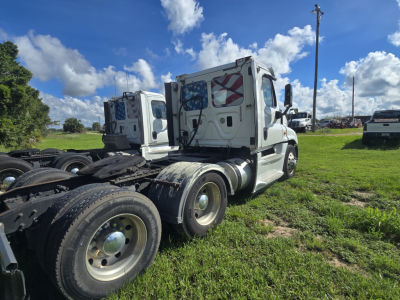 Truck chassis and cab of a 2017 Freightliner Cascadia 125 day cab, white with American flag decals on the rear doors, showing rear triple axle and fifth wheel area, clear sky and grassy lot.