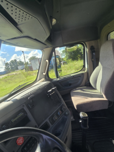 Truck cab interior showing steering wheel, dashboard, gear shifter, and passenger seat; view through side window.