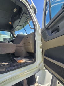 Cab interior view of a Freightliner Cascadia 125 day cab showing driver's seat, dashboard, and open door with step.