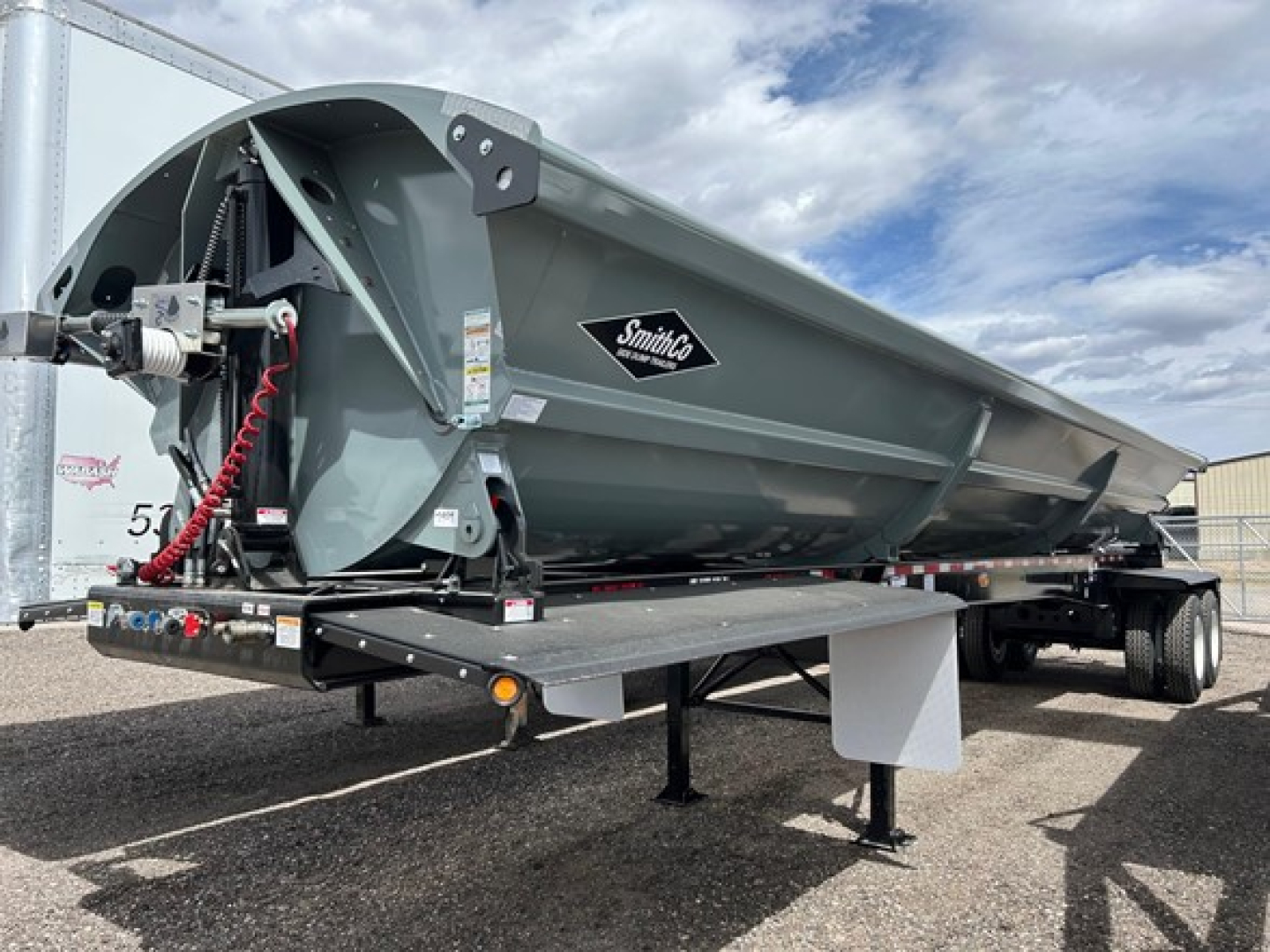 Smithco air ride side dump trailer with light gray body and black frame, front hitch area visible, storage/compartment near wheel, Smithco logo on side.
