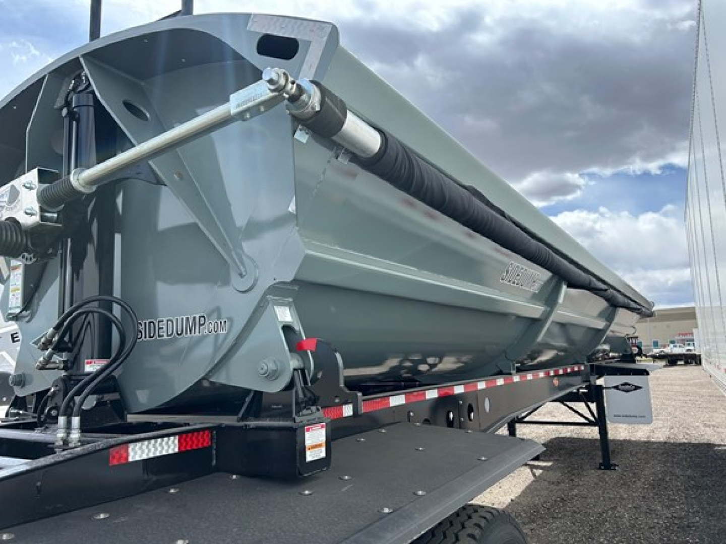Side dump trailer in aluminum gray showing hydraulic cylinder and hydraulic line on a Smithco Air Ride Side Dump with display text 'SIDEDUMP.COM' visible on the hopper.
