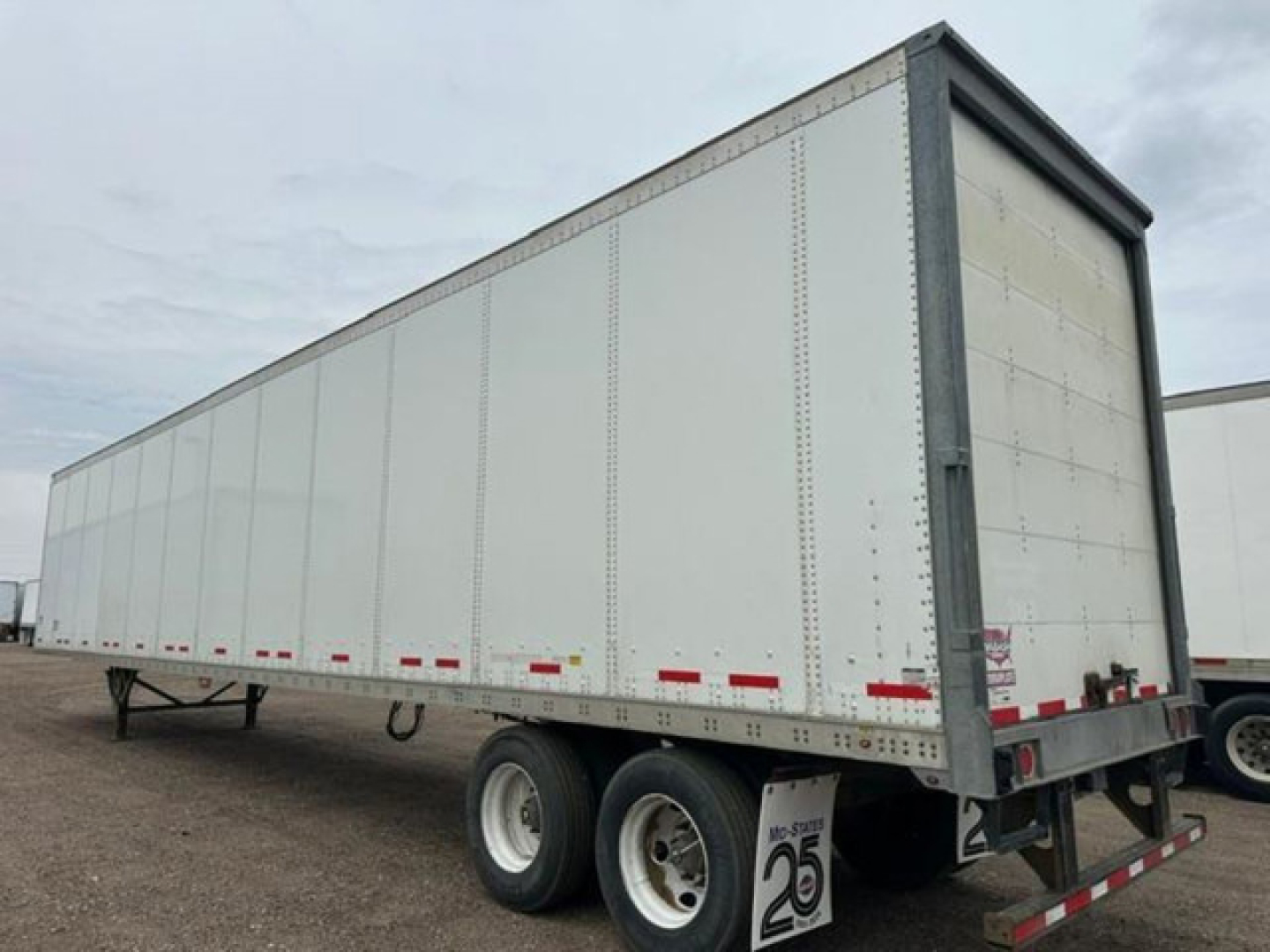 Trailer side view showing a white Wabash van trailer with dual rear axles, no visible branding, and reflective red-and-white tape along the bottom edge.