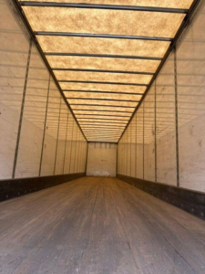 Trailer interior showing wooden floor and side walls with overhead lighting panels; empty cargo space from rear to front of a long box trailer