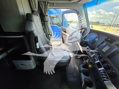 Interior view of a Kenworth truck cab showing driver seat, steering wheel, dashboard, and center console with multiple control panels. Outside window shows blue truck and parking lot.