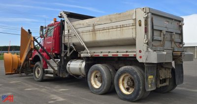Dump truck side view showing a red cab, large aluminum dump bed with tailgate, snow plow attachment, and dual rear axles, weathered surface around the dump bed.