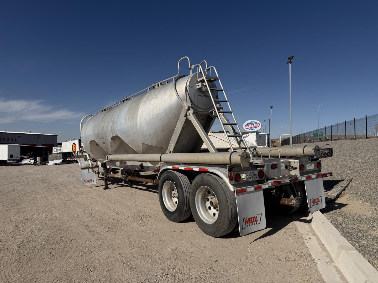 Pneumatic dry bulk trailer in a gravel lot, rear and side views, tank with ladder, dual rear axles, rear bumper lights, Heil branding on mud flaps.
