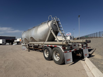 Tank trailer shown in three-quarter rear view, aluminum cylindrical tank, dual rear axles, ladder to top, hoses and piping along chassis, Heil branding visible on mud flaps.