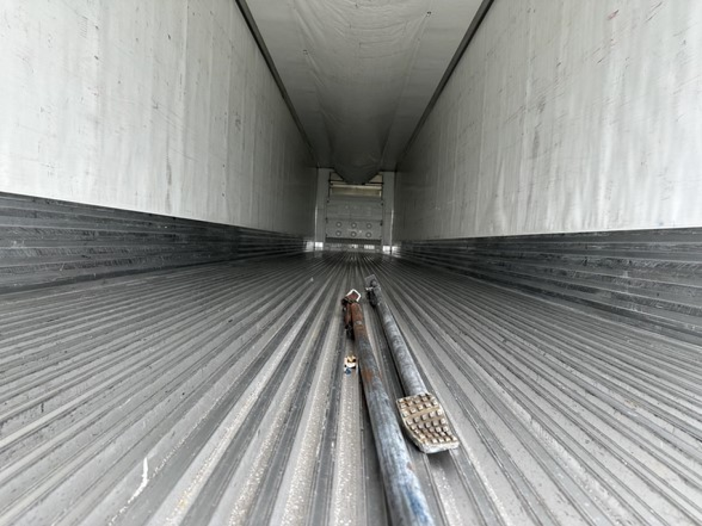 Trailer interior of a 2020 Utility 3000R Reefer showing corrugated metal floor with a long center hinge and two metal rods laid along the floor, perspective toward the rear door.