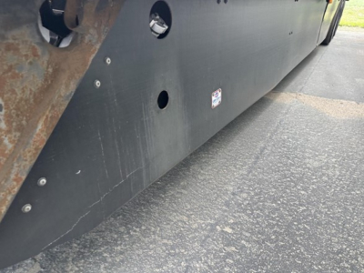 Long side of a utility trailer with a black panel and rusted steel corner. Visible are several circular cutouts and a small sticker on the side. Ground asphalt beneath.