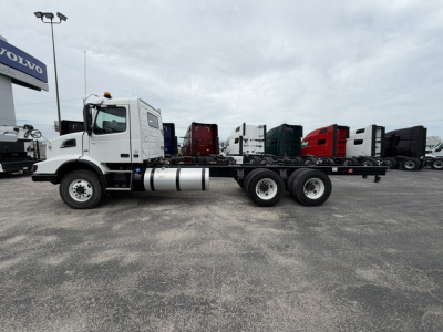 Truck cab and chassis showing Volvo VHD86B300 with long wheelbase and dual rear axles, white cab, empty flatbed, Volvo dealer lot in background.