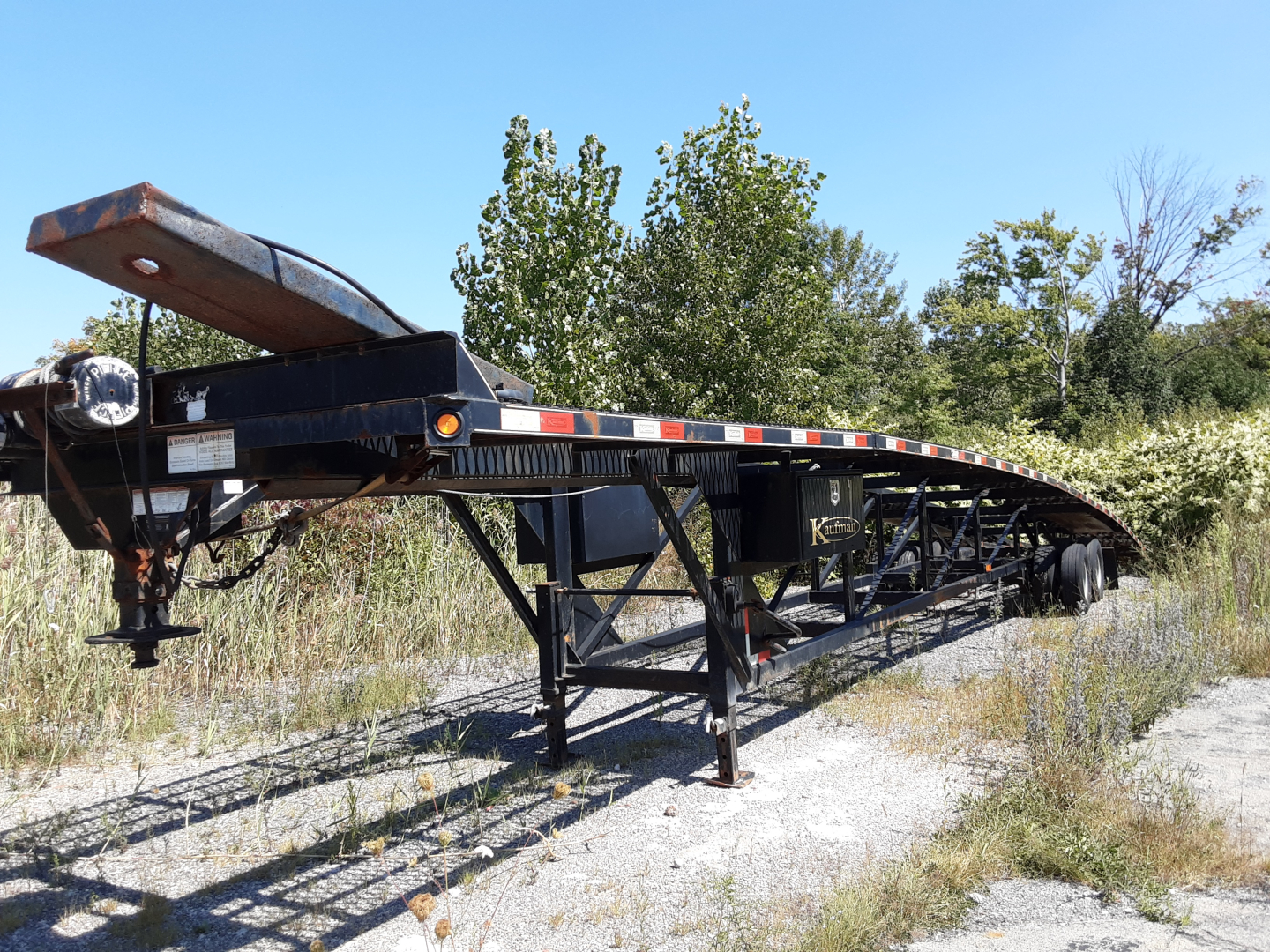 Trailer showing Kaufman 3 car hauler trailer with black frame and tandem wheels parked on gravel.