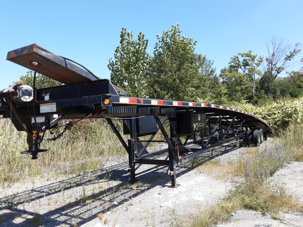 Trailer showing Kaufman 3 car hauler trailer with black frame and tandem wheels parked on gravel.