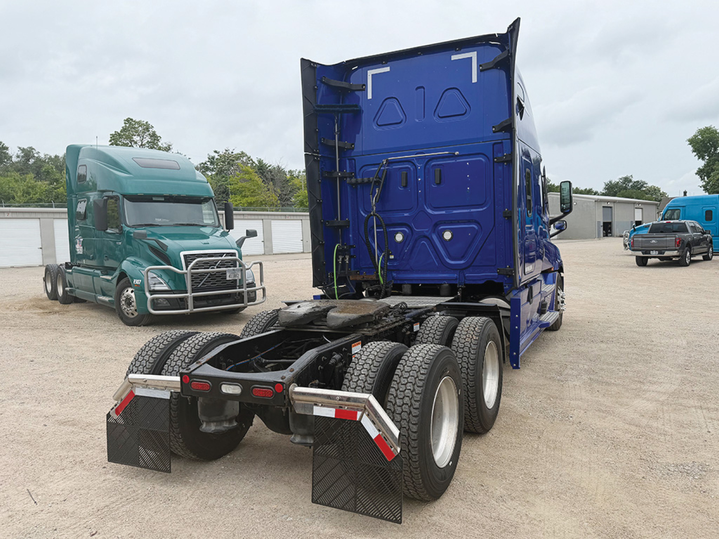 Truck tractor rear view showing blue cab with open sleeper area and dual rear axles, fifth wheel and mud flaps.