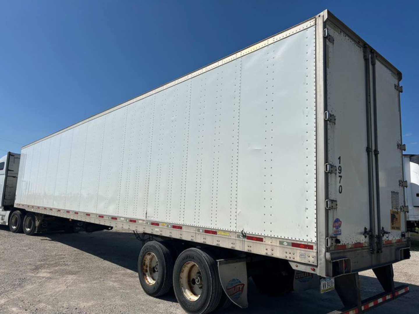 Trailer side and rear view of a white utility van trailer with triple axle wheels, showing riveted panels and rear doors.