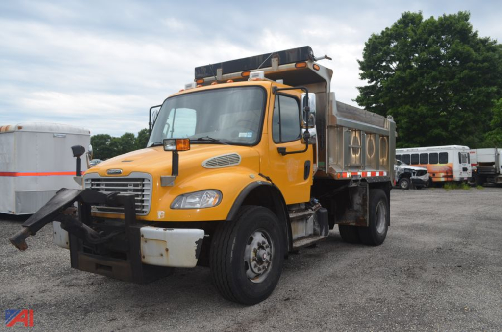 Dump truck in yellow Freightliner M2 106 with dump bed and front bumper guard, side mirrors, and roof lights visible in a lot.