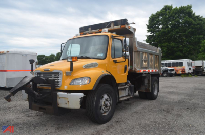 Dump truck in yellow Freightliner M2 106 with dump bed and front bumper guard, side mirrors, and roof lights visible in a lot.