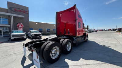 Kenworth T680 tractor in a dealership lot, red cab, dual rear axles, viewed from rear-side angle showing fuel tanks and wheels.