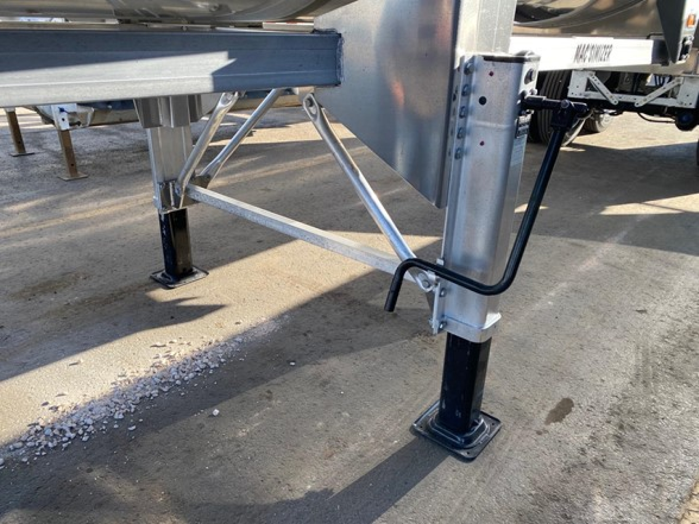 Trailer leveling jack and structural supports visible under a MacMacsimiser 1/2 Round dump trailer, showing metal frame, side stabilization leg, and ground contact pad.