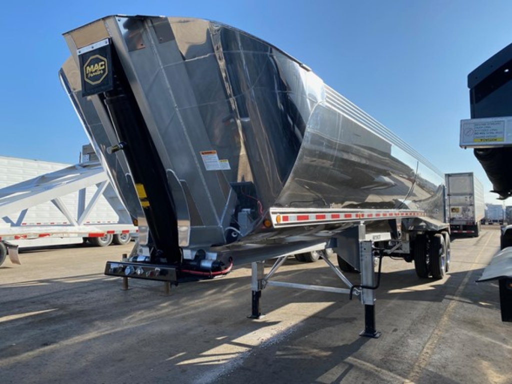 Mac Macsimiser 1/2 Round dump trailer showing polished steel body and rear hitch. Visible: Mac logo, trailer bed, support legs, red-white reflective tape.