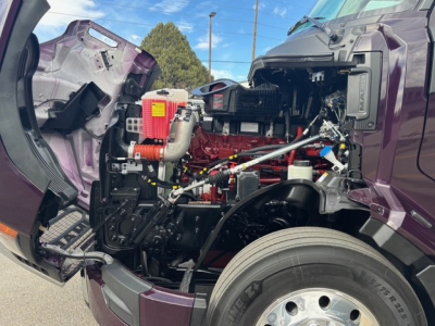 Truck engine bay of a 2026 Mack PR64T76H showing red engine block, intake, hoses, and surrounding components; cab tilted with purple exterior.