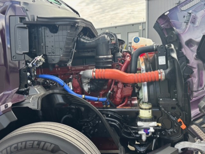 Engine bay of a Mack PR64T76H with exposed red engine, blue and orange hoses, and surrounding purple cab panels.