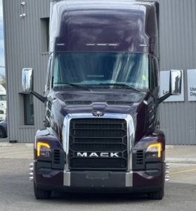 Truck front view of a maroon 2026 Mack PR64T76H with large grille, illuminated headlights, and chrome mirrors.